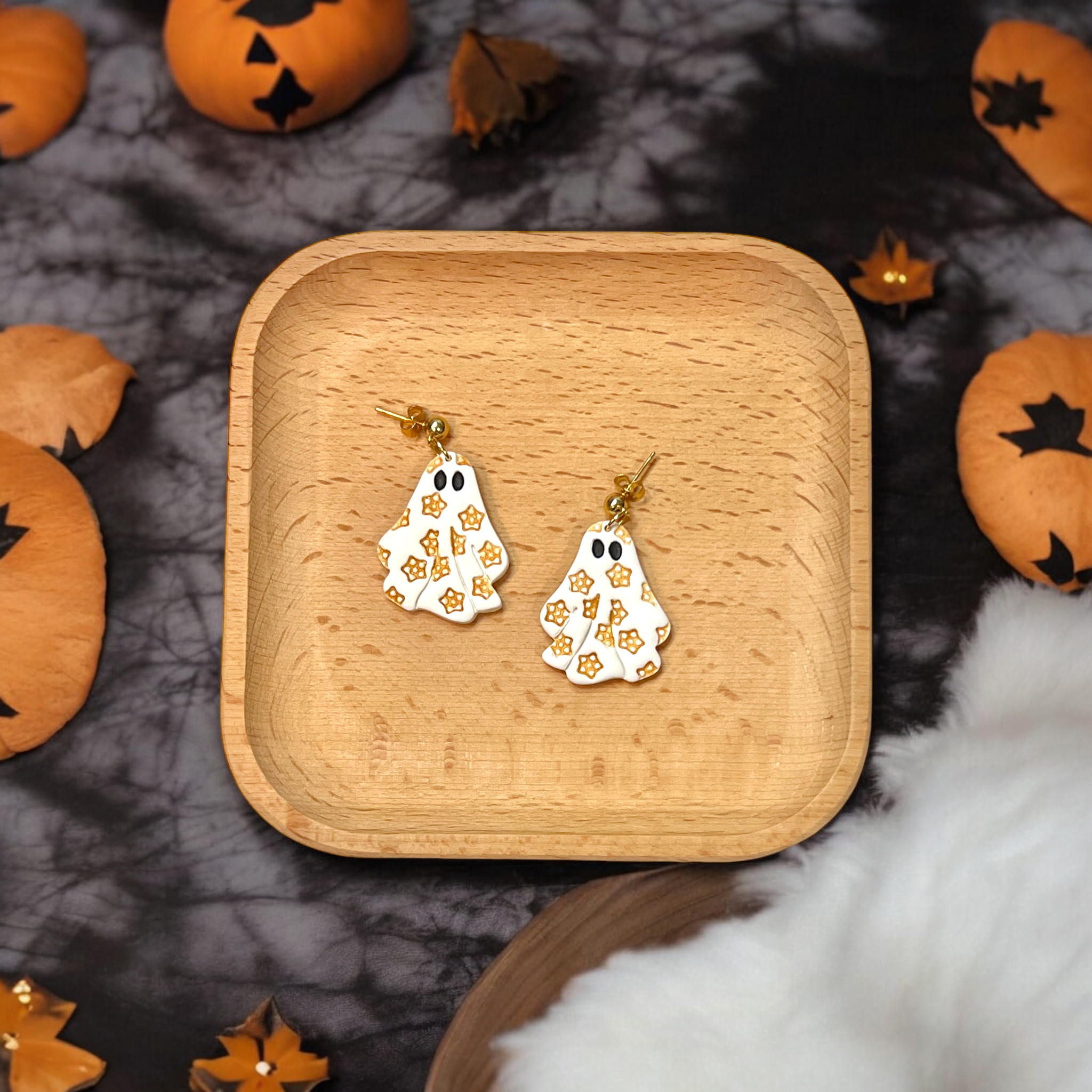 A pair of polymer clay ghost-shaped dangle earrings with golden star detailing, displayed on a wooden tray, surrounded by Halloween decorations.