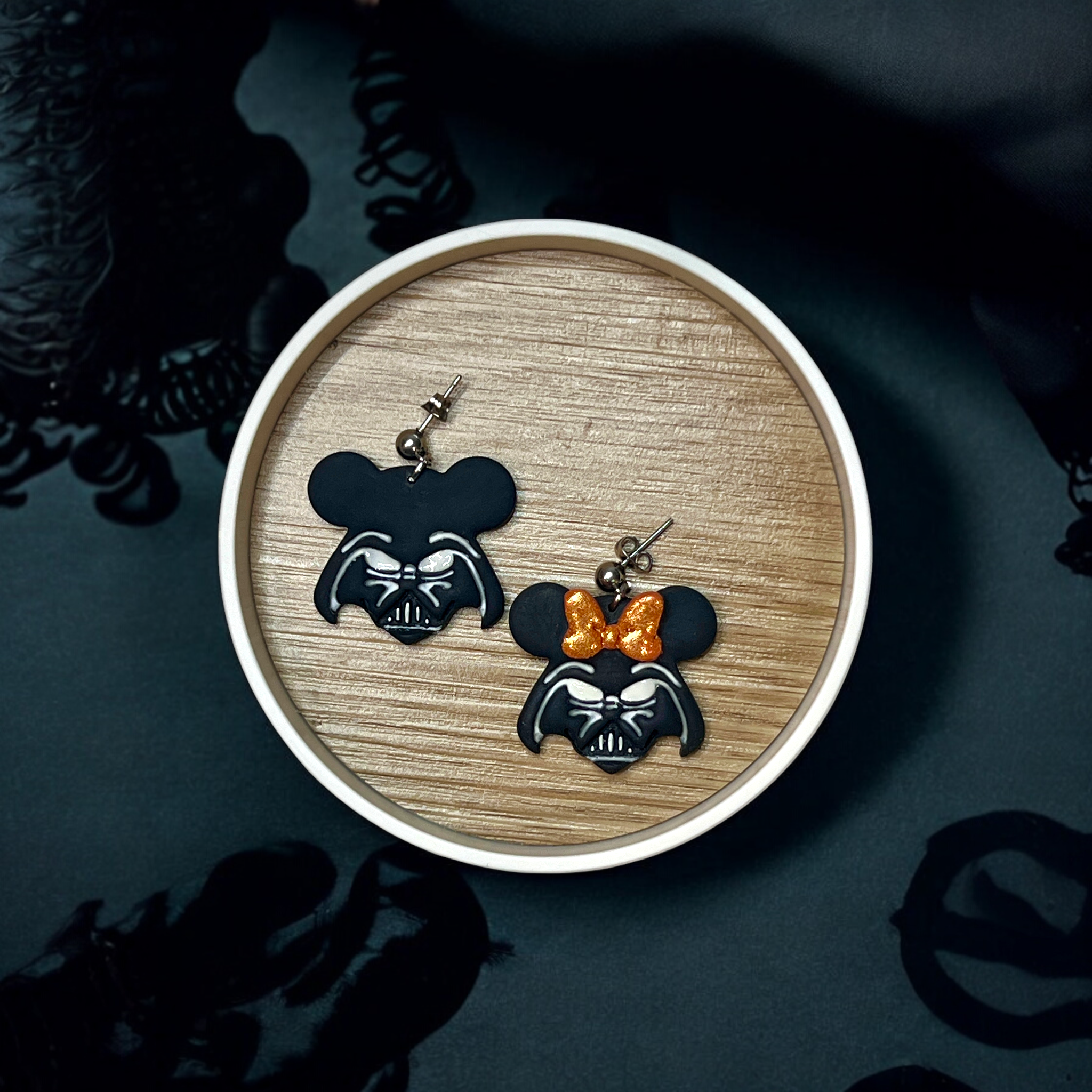 Close-up of Star Wars-themed polymer clay dangle earrings with a Darth Vader and mouse ear design, one adorned with an orange glitter bow, placed on a wooden tray with a dark backdrop.