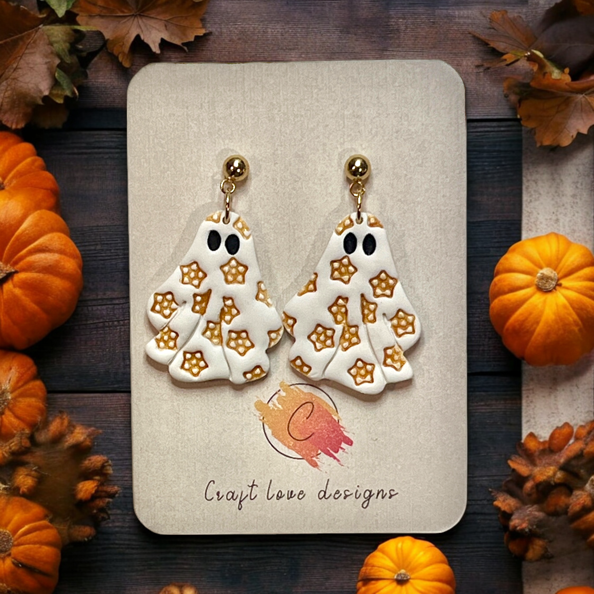 Close-up of polymer clay ghost earrings with golden star detailing, displayed on a Halloween-themed background with pumpkins and autumn leaves.