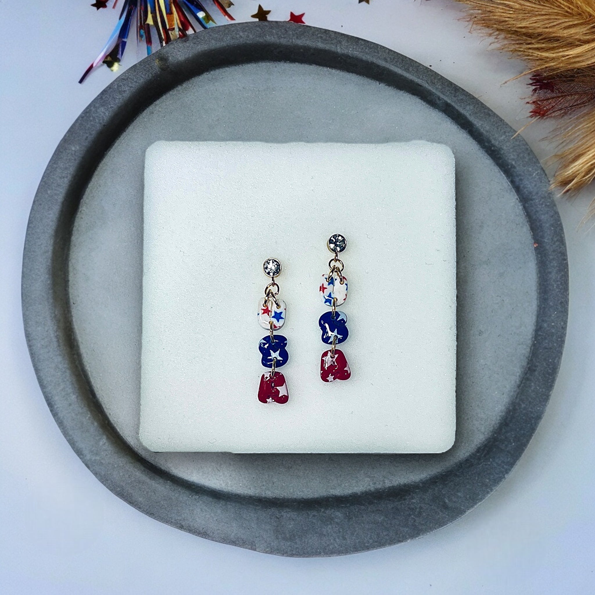 Patriotic red, white, and blue star earrings with gold hardware and rhinestone studs, displayed on a white cushion with festive decor.